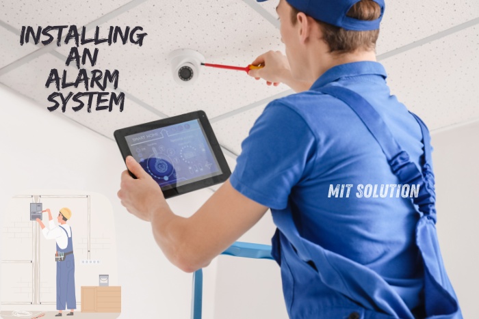 A technician installing an alarm system in a ceiling, using a tablet for configuration, representing the professional security services provided by MIT Solution in Dindigul, Tamil Nadu. The caption 'Installing an Alarm System' highlights MIT Solution's expertise in modern alarm system installations for enhanced safety